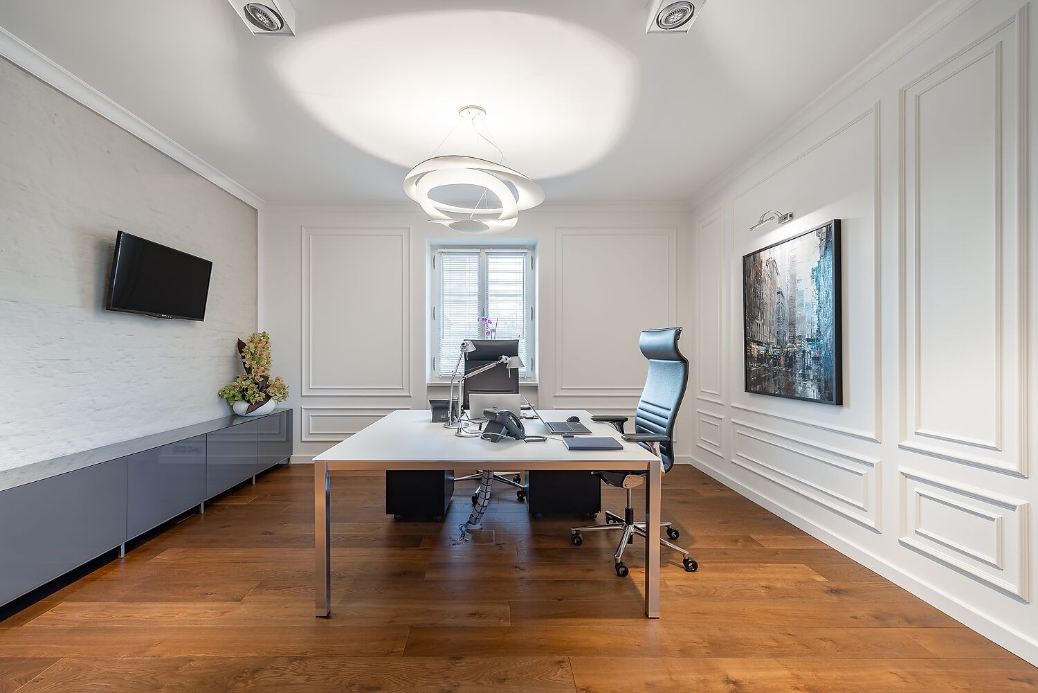 Nowoczesne biuro z białymi ścianami zdobionymi sztukaterią, drewnianą podłogą, szarym kredensem, wiszącym telewizorem, białym biurkiem z laptopem i czarnym krzesłem biurowym