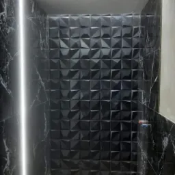 Wnętrze łazienki z czarnymi płytkami imitującymi marmur na ścianach i trójwymiarowymi płytkami na jednej ze ścian, oświetlone listwą LED i surową żarówką.