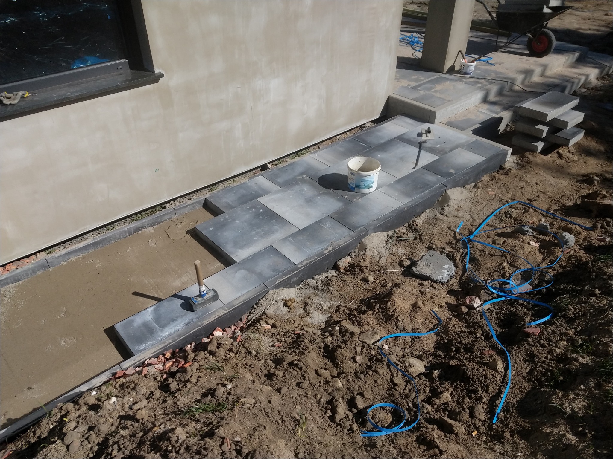 Fragment układanej ścieżki z szarych płyt betonowych przy budynku, widoczne narzędzia, wiadro i niebieski przewód na ziemi.