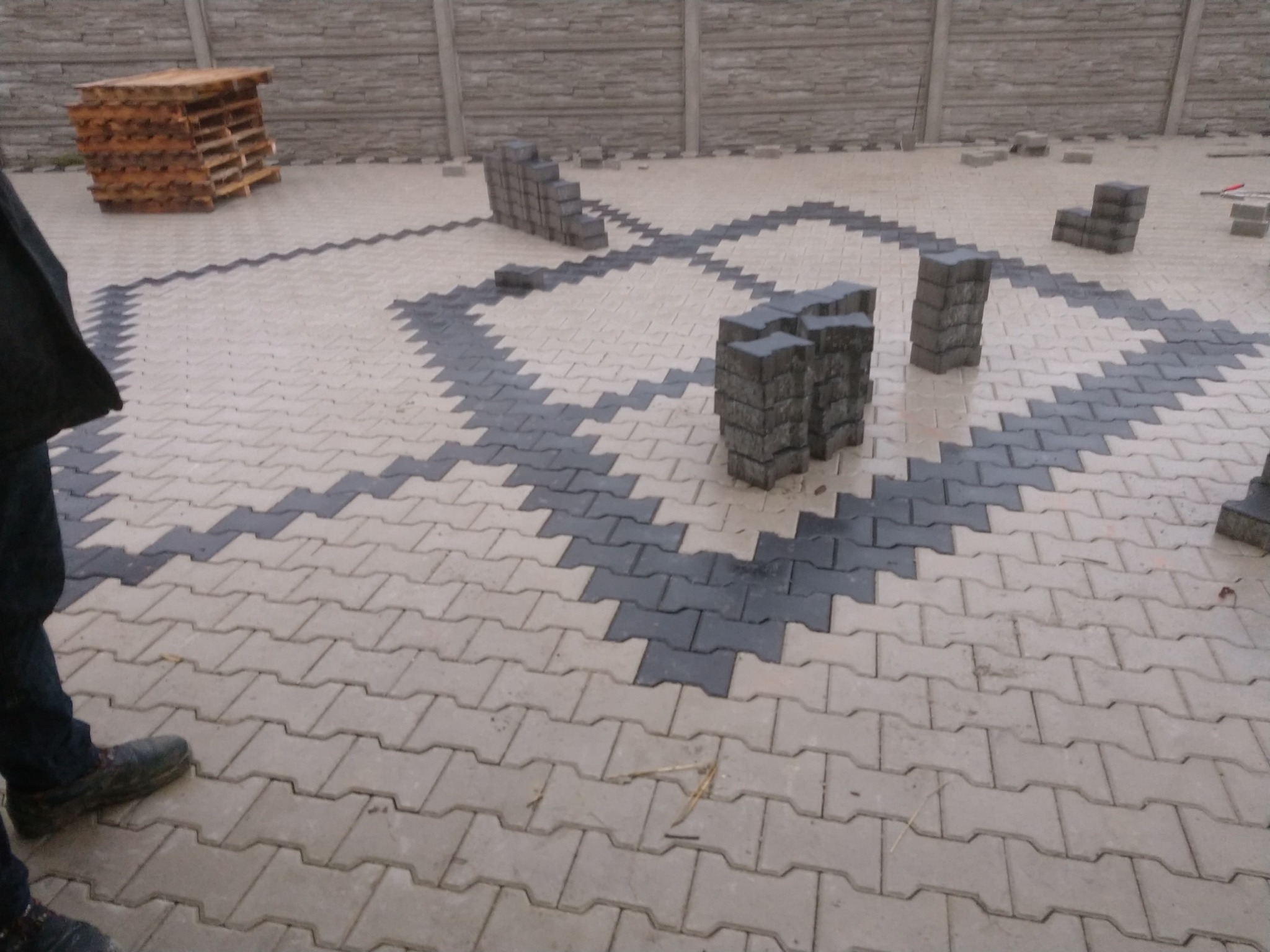 Układanie kostki brukowej w geometryczny wzór z użyciem szarej i beżowej kostki, widoczne palety z kostką w tle.