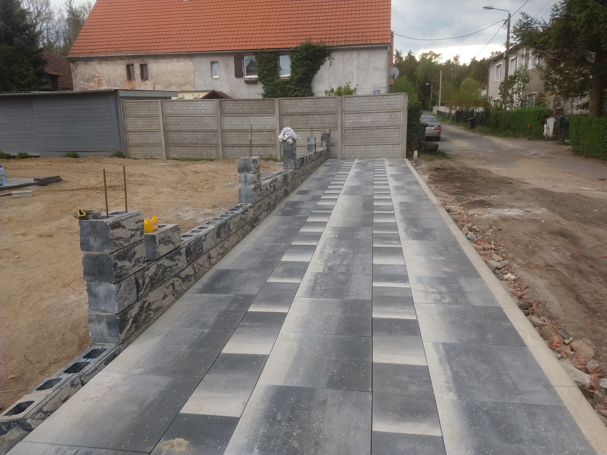 Częściowo ułożona nawierzchnia z szarych płyt betonowych, obok mur z bloczków betonowych w trakcie budowy, narzędzia na murze, dom w tle.