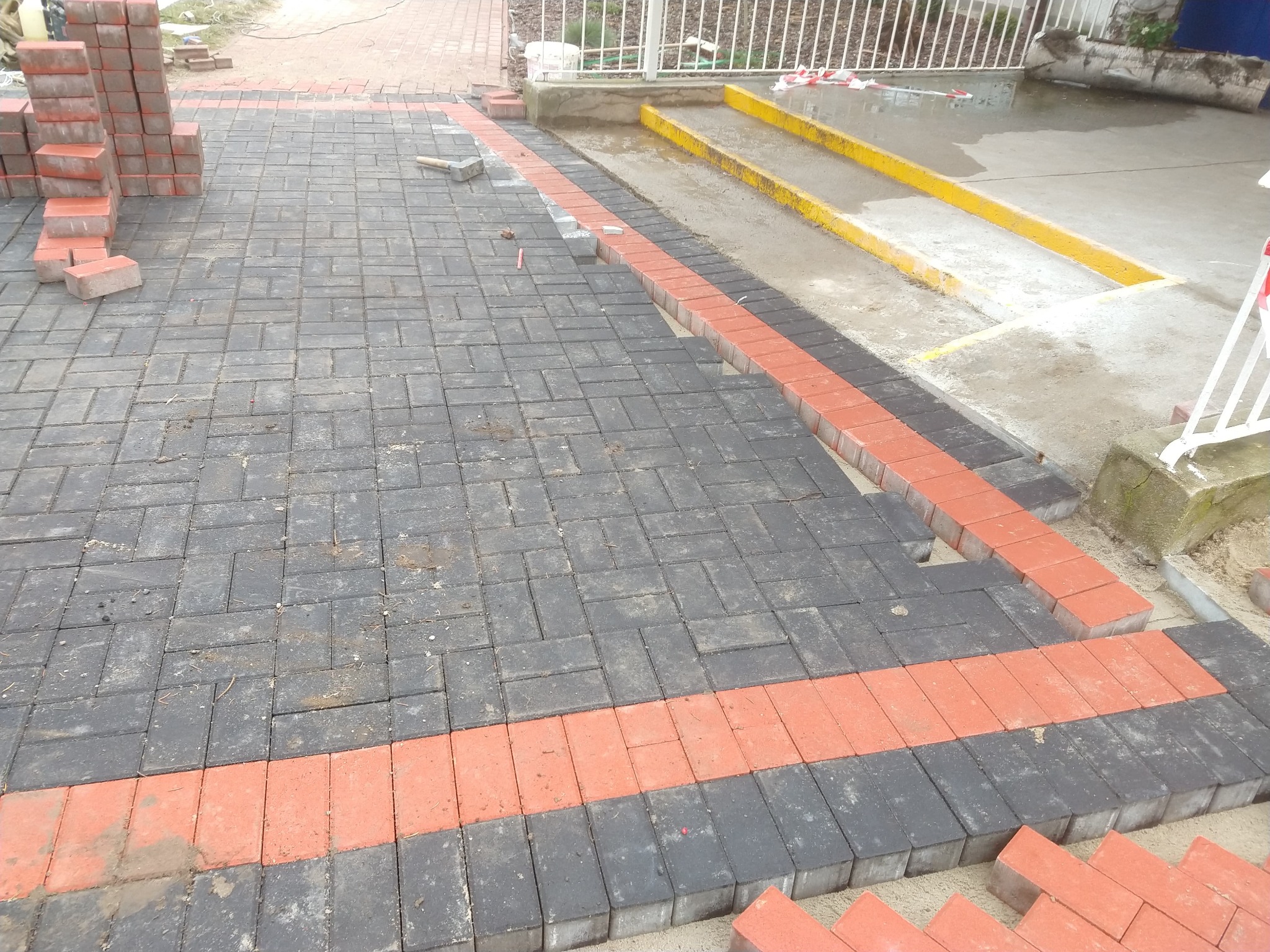 Częściowo ułożona nawierzchnia z kostki brukowej w kolorze grafitowym z bordiurą z czerwonej kostki; widoczny stos kostki, młotek brukarski i elementy zabezpieczenia terenu.