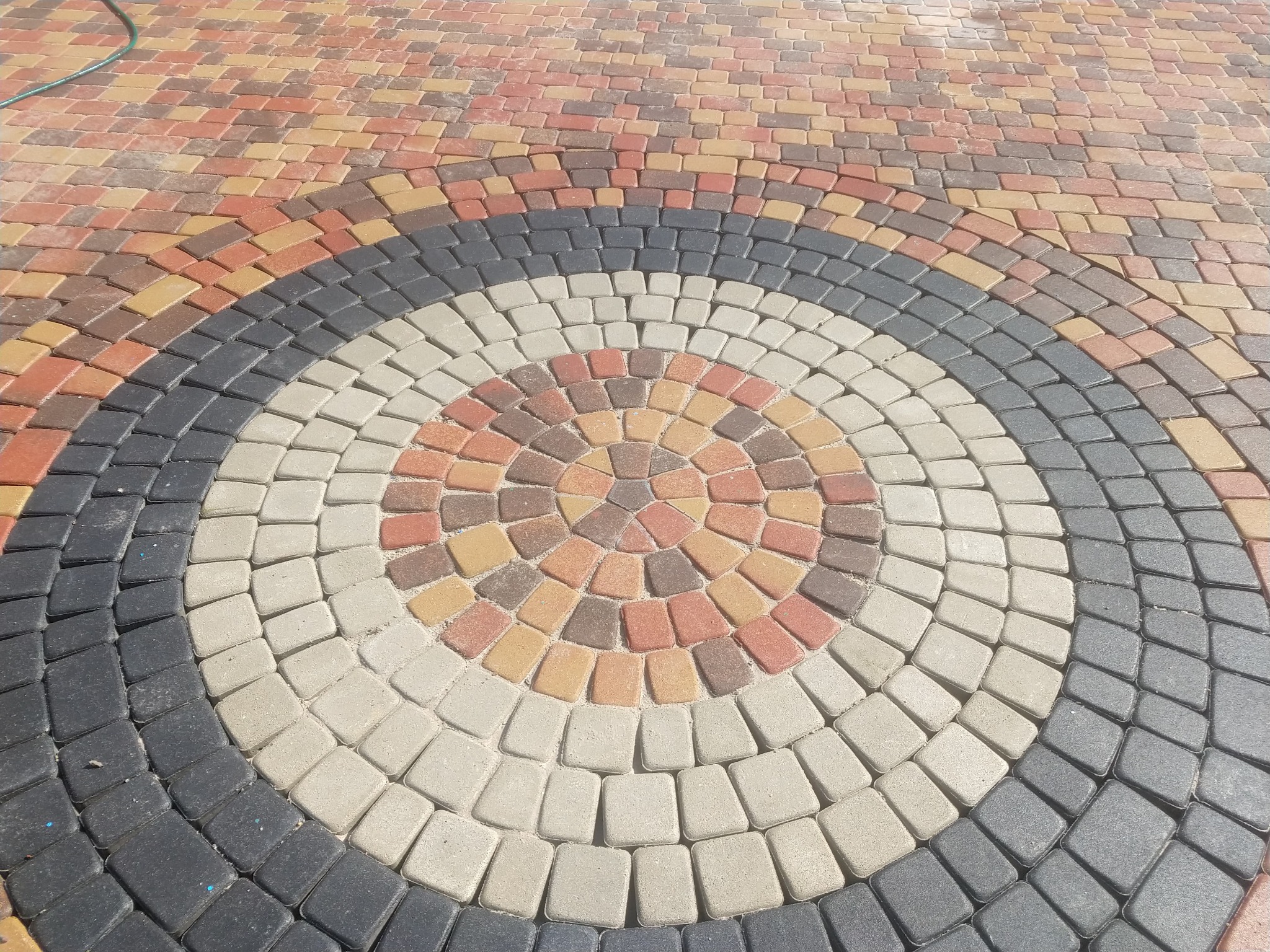 Kolorowa kostka brukowa ułożona w koncentryczny wzór z dominującymi odcieniami czerwieni, brązu, szarości i beżu, tworząca dekoracyjny okrąg na nawierzchni.