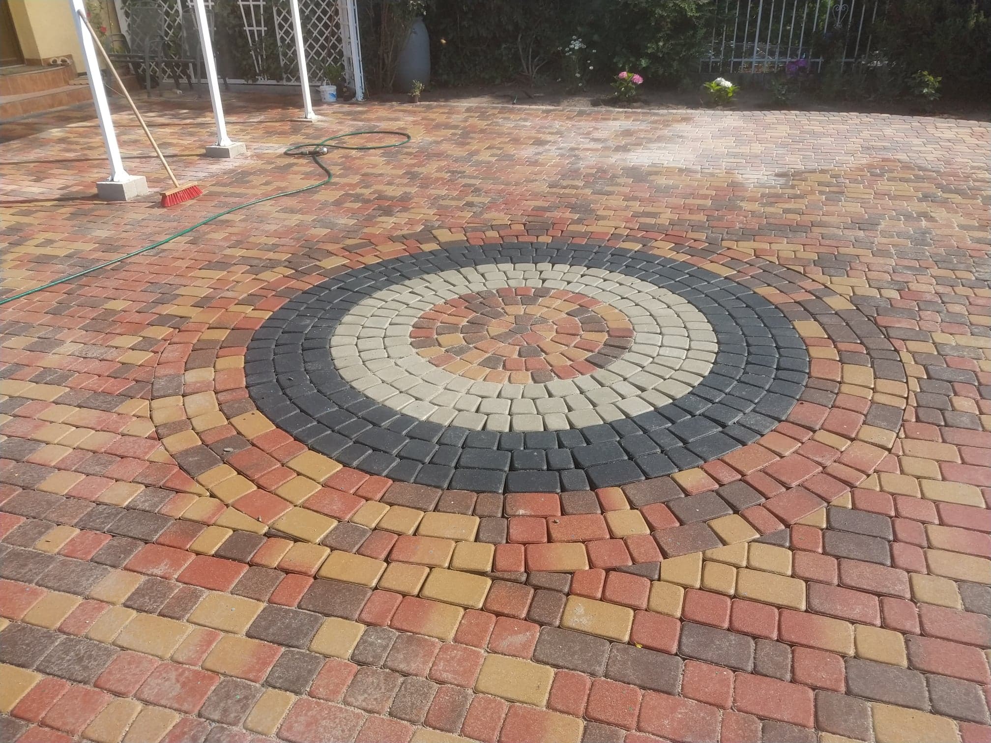 Kolorowa nawierzchnia z kostki brukowej ułożona w okrągły wzór z elementami czarnymi, beżowymi, pomarańczowymi i czerwonymi; w tle widoczny fragment domu i zieleń.
