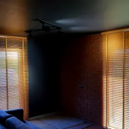 Wnętrze pokoju z drewnianymi żaluzjami poziomymi, widok na okno i ścianę z cegły, oświetlenie punktowe na suficie, fragment niebieskiej sofy.