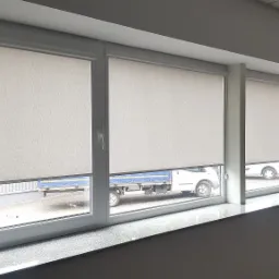 Cztery okna z zamontowanymi, częściowo opuszczonymi roletami w jasnym odcieniu szarości, widok na zaparkowane samochody dostawcze na zewnątrz.