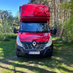 Czerwony Renault Master z wysoką zabudową, zaparkowany na trawie w pobliżu drzew, widok z przodu.