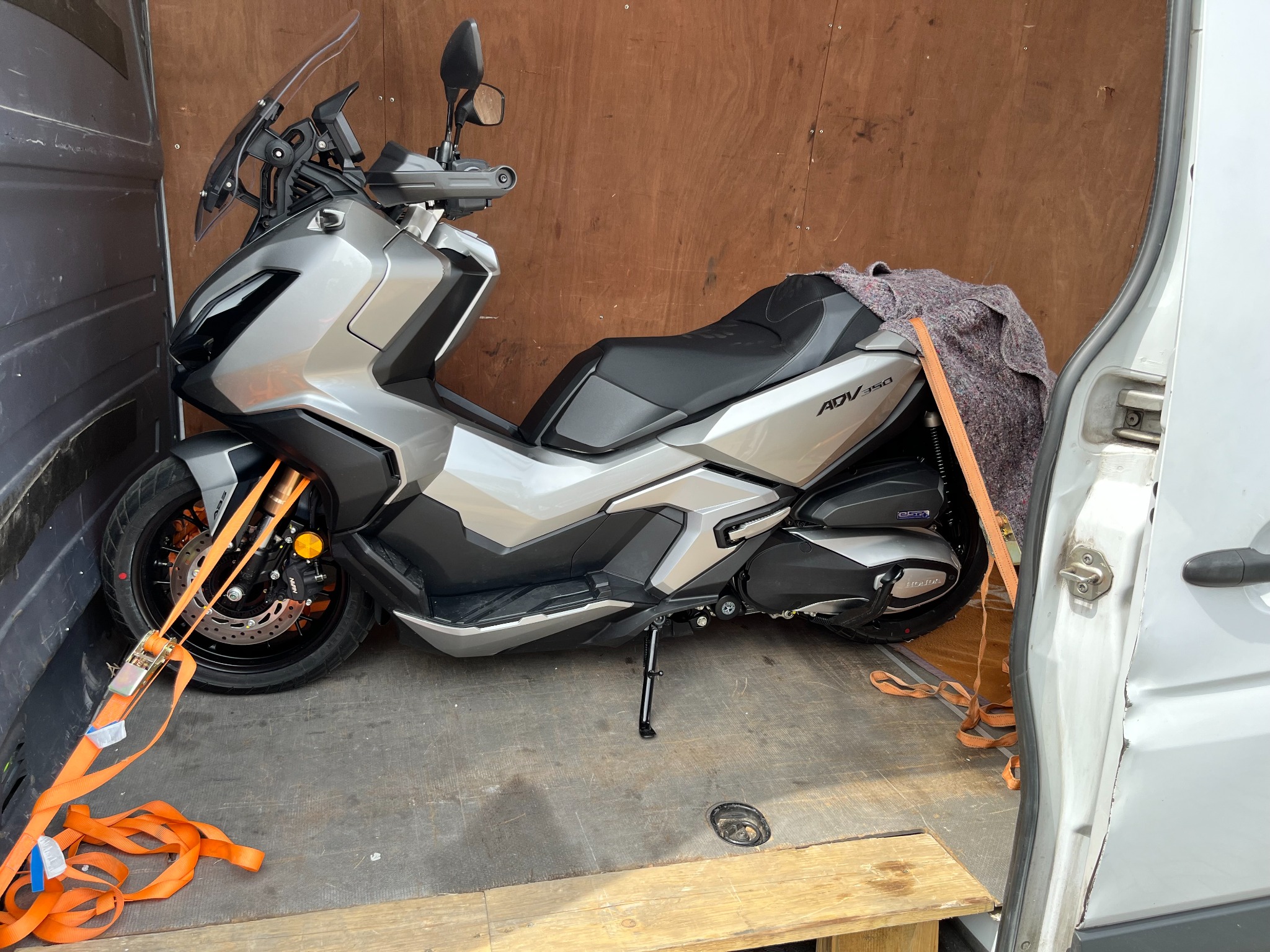 Srebrny motocykl Honda ADV 350 zabezpieczony pomarańczowymi pasami wewnątrz furgonetki z drewnianą ścianą.