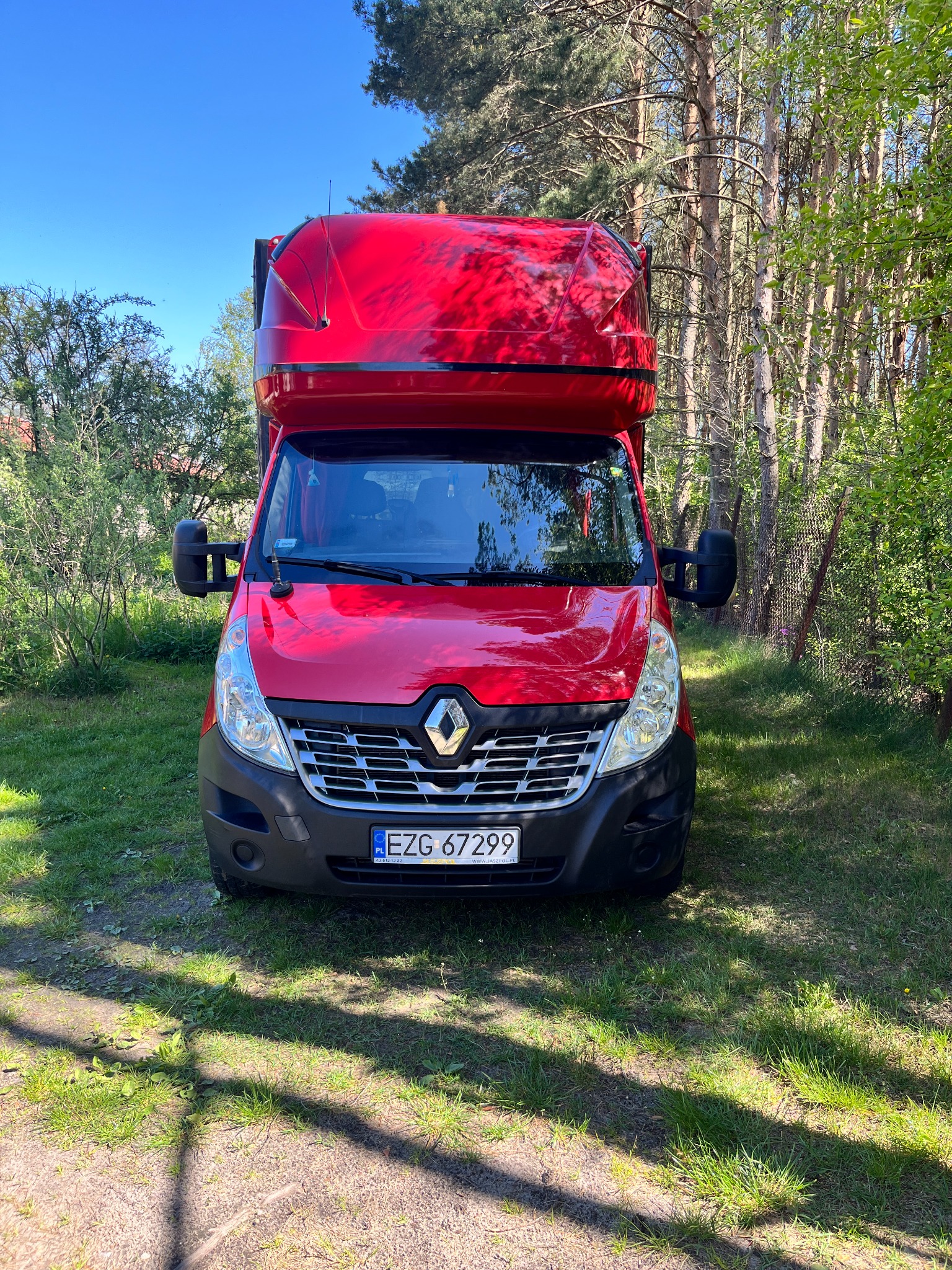 Czerwony Renault Master z wysoką zabudową, zaparkowany na trawie w pobliżu drzew, widok z przodu.