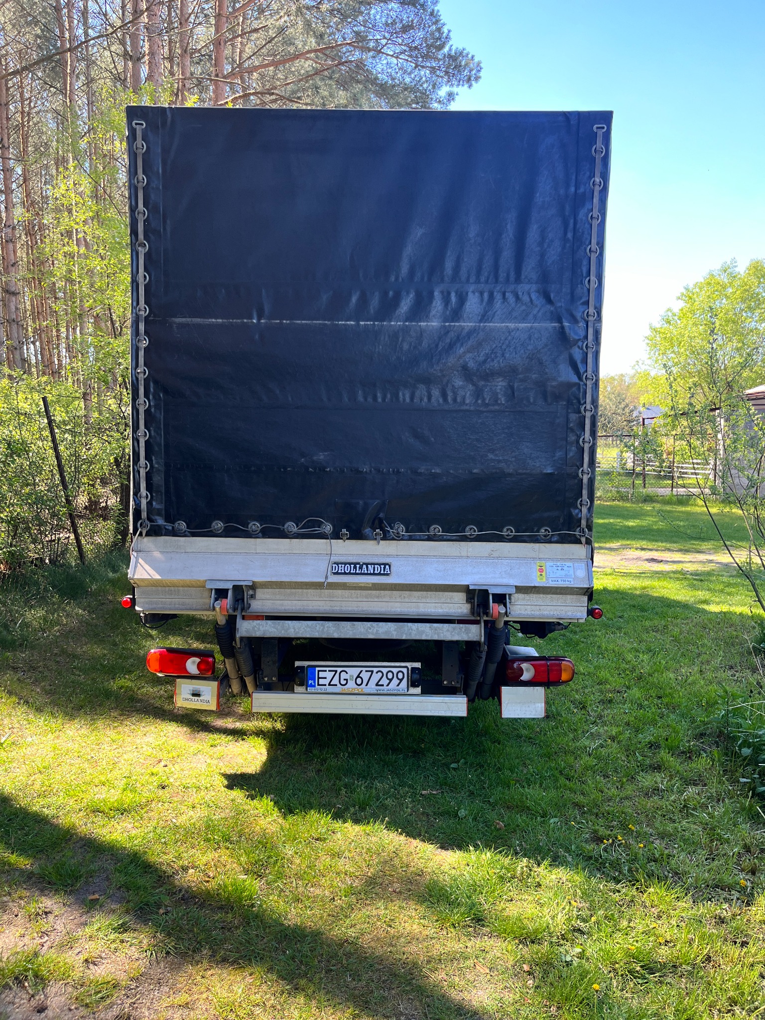 Tył samochodu dostawczego z plandeką i windą załadowczą Dhollandia na tle zielonej trawy i drzew, widoczna tablica rejestracyjna EZG 67299.