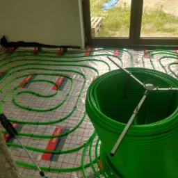 Rozłożone, zielone rury ogrzewania podłogowego na folii izolacyjnej z widocznym oknem i stertą rur w pomieszczeniu w trakcie montażu.