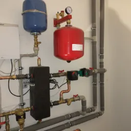 Zmontowana instalacja grzewcza z piecem Vaillant, czerwonym i niebieskim zbiornikiem wyrównawczym, czarnym rozdzielaczem i rurami z szarą izolacją termiczną.