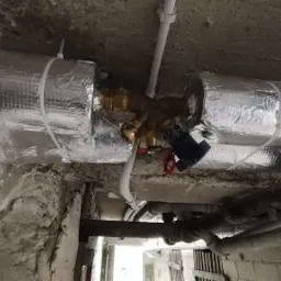 Izolowane rury instalacji wodnej z zaworami i przyłączami, widoczne w surowym pomieszczeniu z betonowym stropem i ścianami.