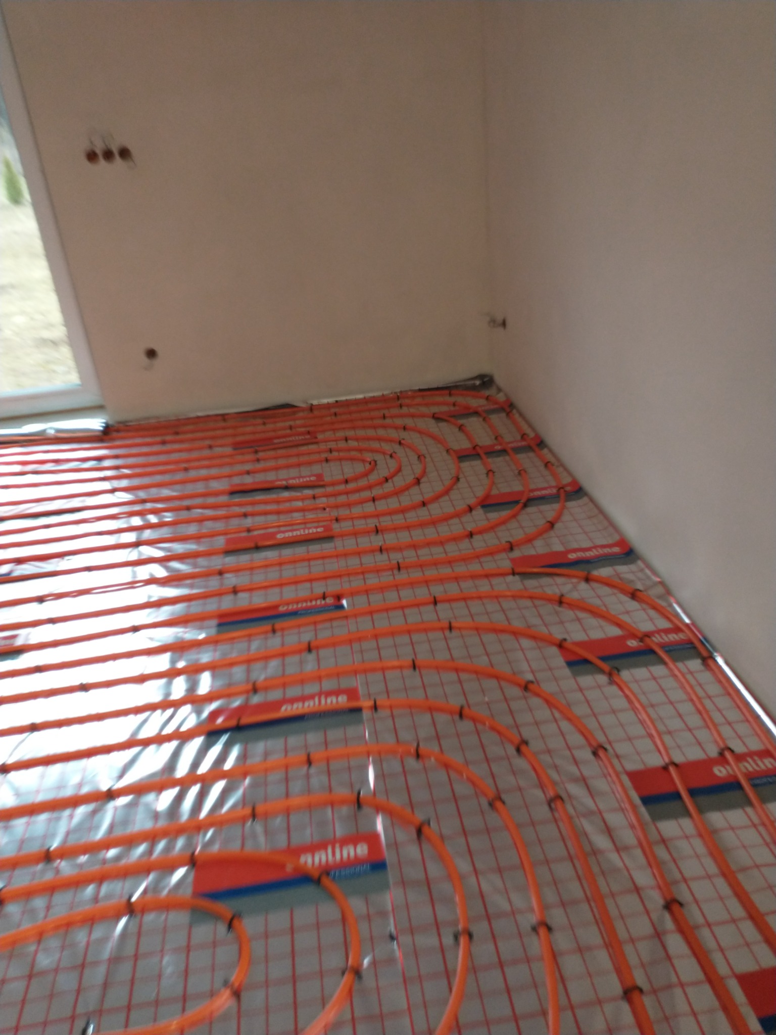 Pomarańczowe rury ogrzewania podłogowego ułożone na folii izolacyjnej z widocznymi uchwytami mocującymi i fragmentem okna w tle.