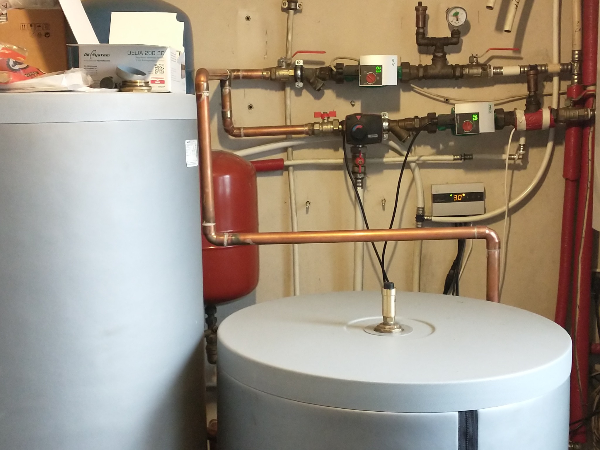 Dwa szare zasobniki na wodę połączone miedzianymi rurami, widoczne zawory, manometry i sterowniki elektroniczne na ścianie, instalacja w kotłowni.