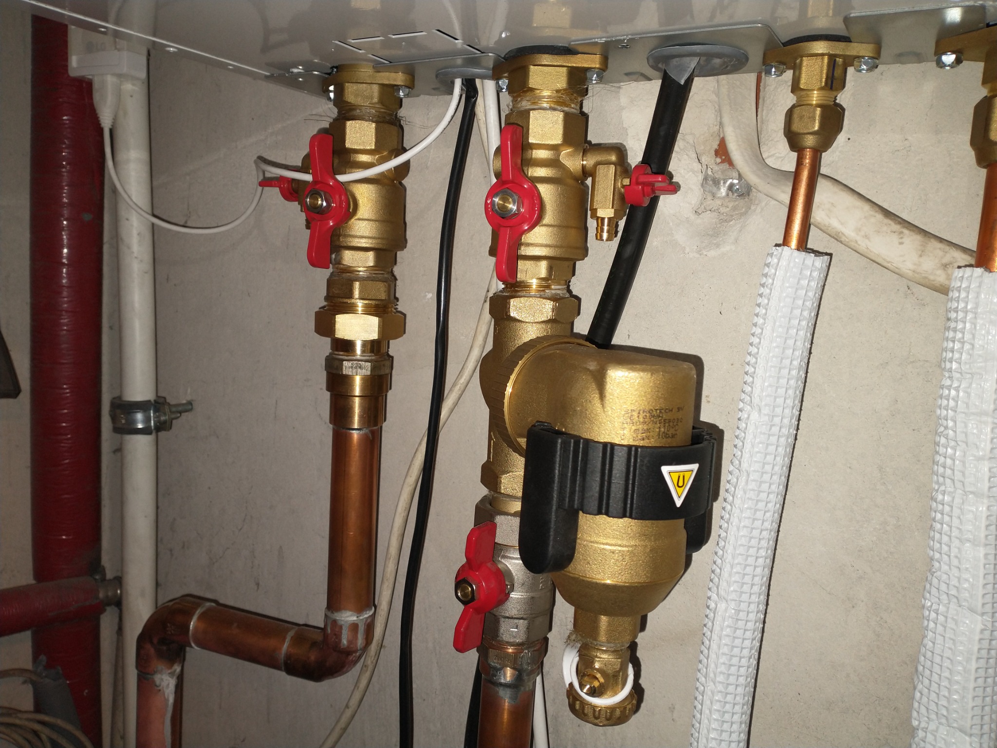 Złote zawory z czerwonymi pokrętłami połączone miedzianymi rurami, element instalacji z filtrem magnetycznym, widoczne białe kable i izolowane rury.
