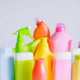 Zestaw kolorowych butelek i pojemnik&oacute;w z płynami do czyszczenia, w pastelowych odcieniach, na jasnym tle, z zielonym spryskiwaczem na pierwszym planie.