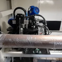 Maszyna do cięcia materiałów w roli, zabezpieczona folią, widoczne elementy mechaniczne i silniki.