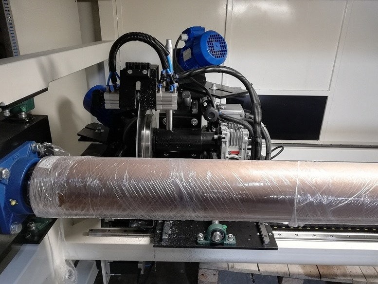 Maszyna do cięcia materiałów w roli, zabezpieczona folią, widoczne elementy mechaniczne i silniki.