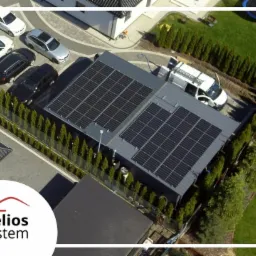 Widok z góry na instalację fotowoltaiczną na dachu garażu samochodowego z białym samochodem serwisowym obok, na tle zadbanego ogrodu i kilku zaparkowanych samochodów.