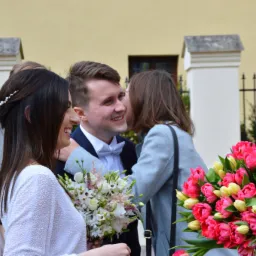 Uśmiechnięta para młoda w otoczeniu gości z bukietami kwiatów podczas ceremonii ślubnej na zewnątrz budynku.