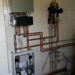 Kocioł gazowy kondensacyjny z otwartą szafką rozdzielczą, widoczne miedziane rury i armatura, na ścianie z płytek.