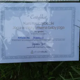 Certyfikat ukończenia kursu nauczycielskiego Joga w ciąży i mama baby joga na 100 godzin, przyznany Magdalenie Juchniewicz, podpisany przez Agnieszkę Berę z Shakti Doula Copenhagen, leżący na trawie.
