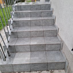 Mariusz ŚWIĄTEK - Zewnętrzne schody wejściowe wyłożone szarą, kwadratową płytką z delikatnym wzorem, zabezpieczone metalową, pomalowaną na szaro barierką; widoczne niewielkie zabrudzenia na krawędziach płytek.