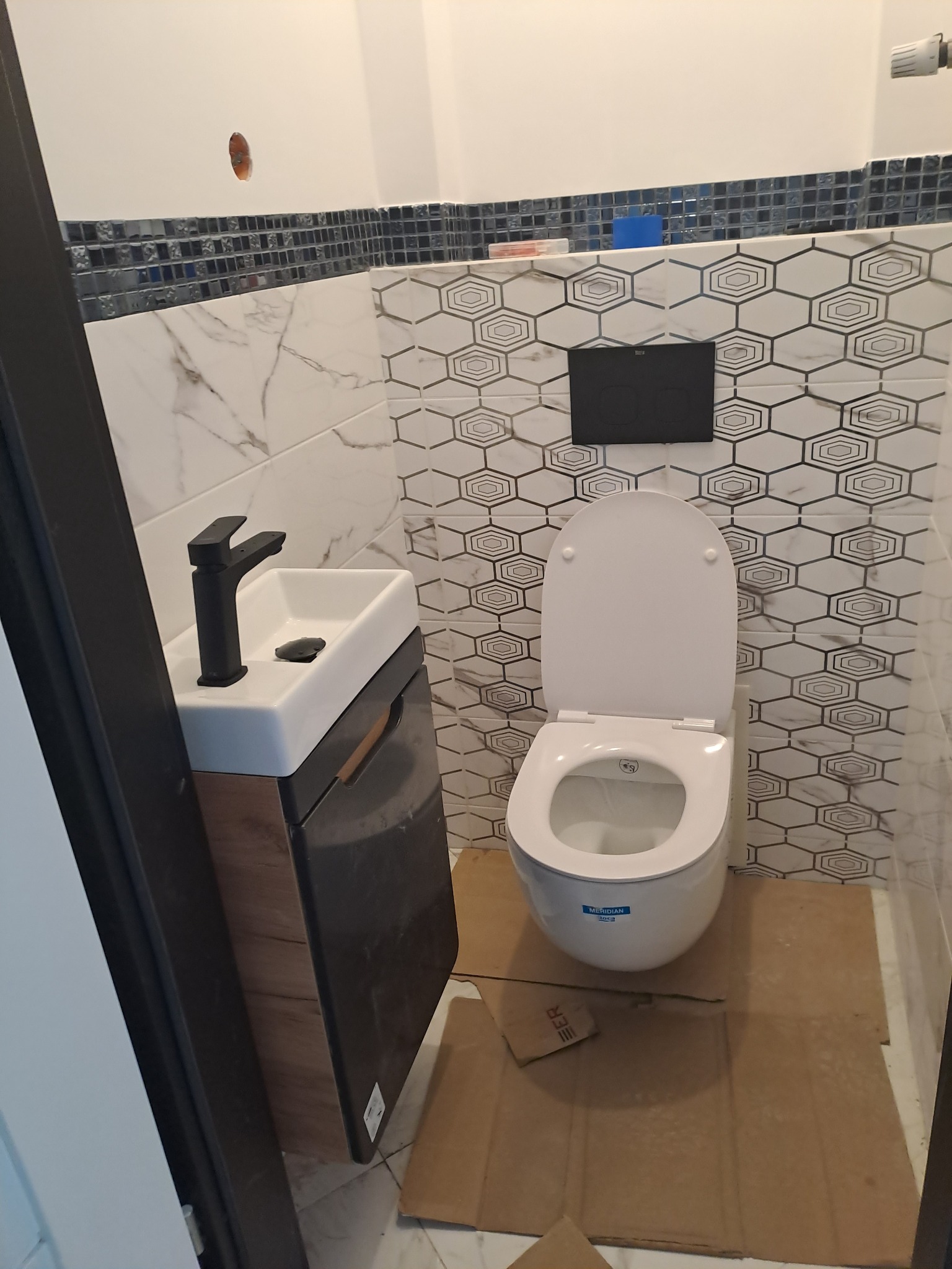 Nowoczesna łazienka w trakcie wykończenia: biała umywalka nablatowa z czarną baterią, wisząca toaleta z deską, mozaika z geometrycznym wzorem na ścianie, zabezpieczona podłoga kartonem.