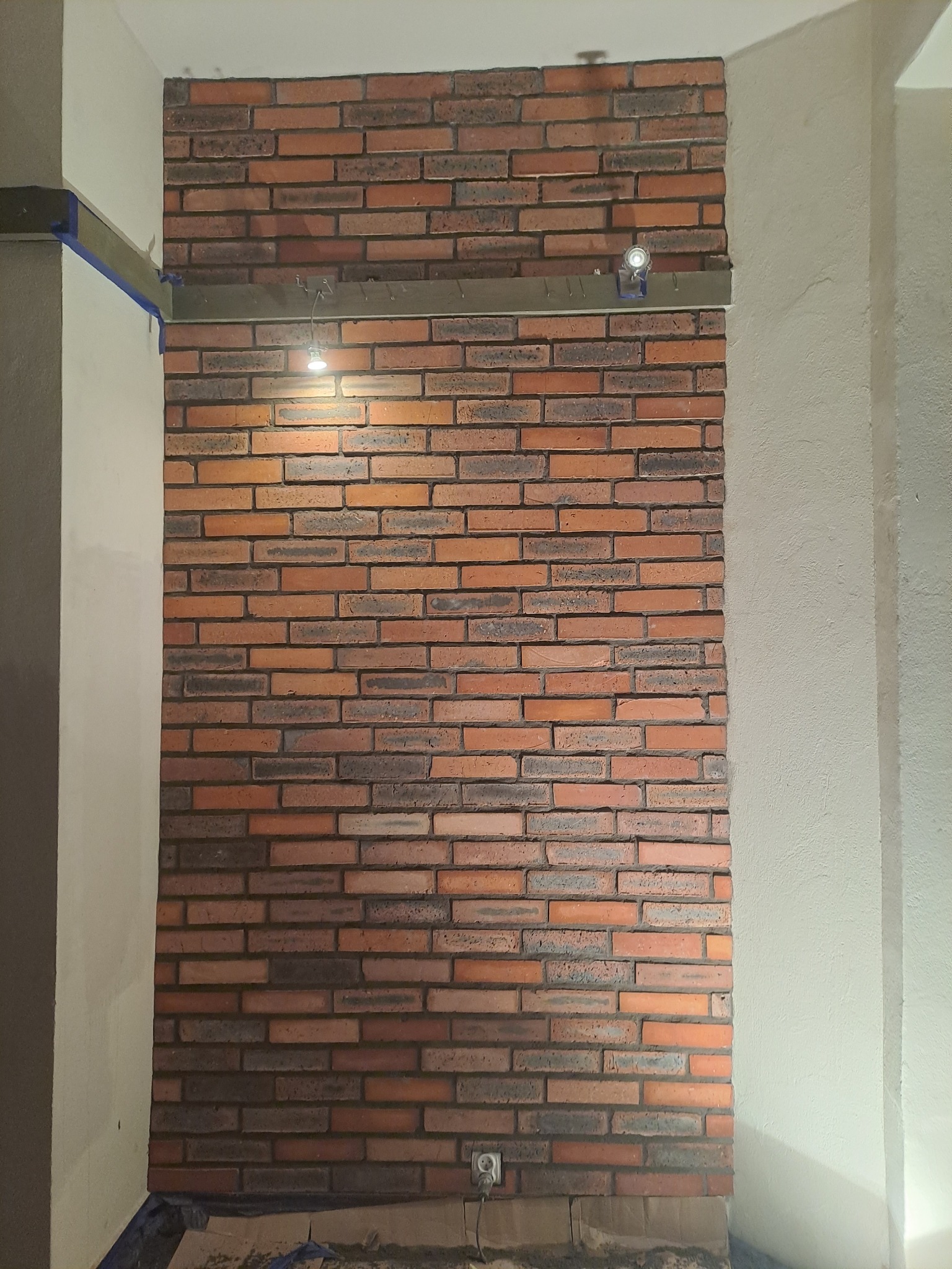Ściana z cegły klinkierowej wewnątrz pomieszczenia, oświetlona punktowo, z widocznym fragmentem instalacji elektrycznej i zabezpieczeniem narożnika taśmą malarską.