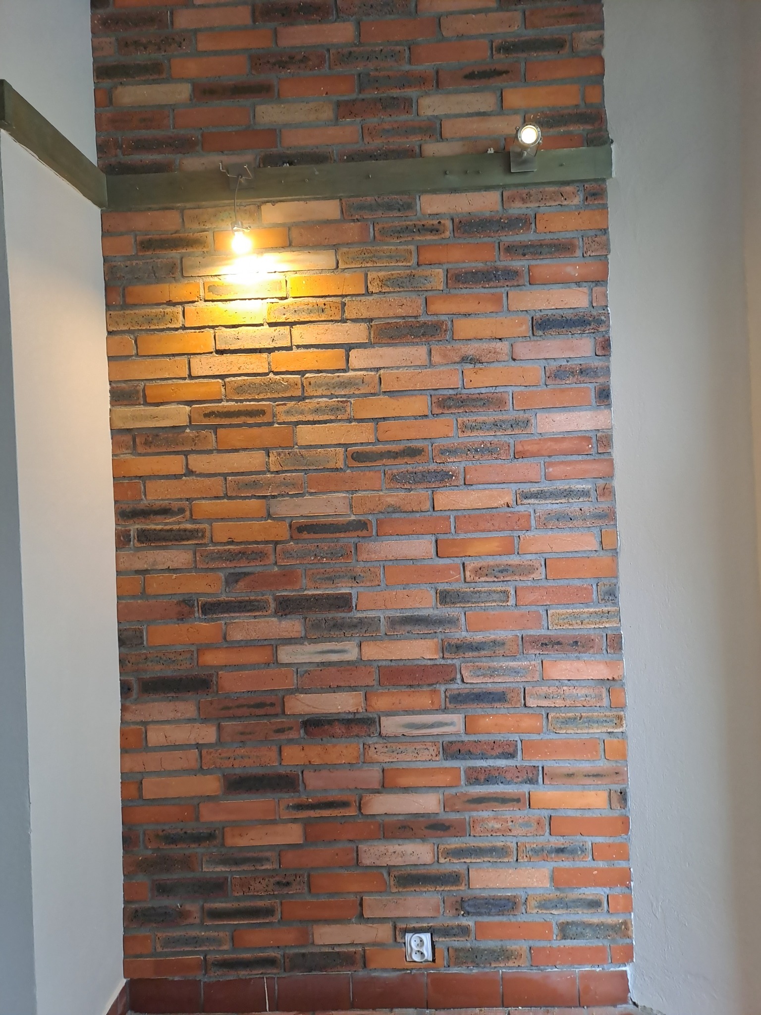 Ściana z cegły klinkierowej w odcieniach czerwieni i brązu, oświetlona dwoma lampami punktowymi, z widocznym gniazdkiem elektrycznym u dołu.