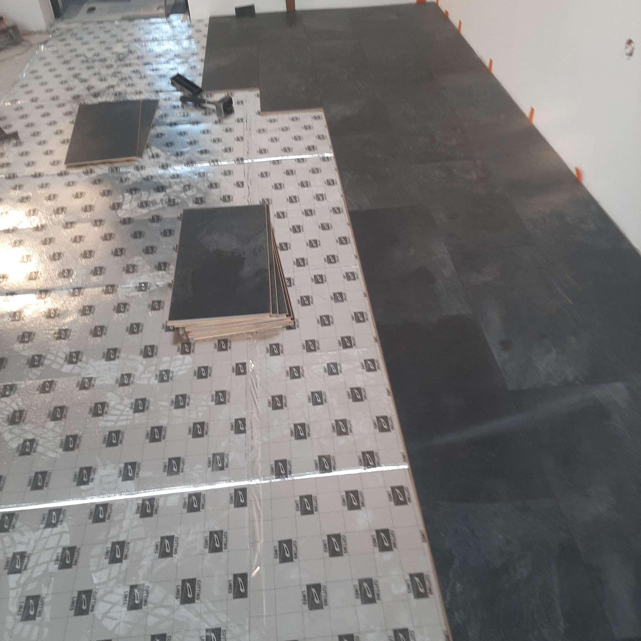 Układanie ciemnych płytek podłogowych na folii z nadrukiem, widoczne narzędzie do docinania i pomarańczowe kliny dystansowe przy ścianie.