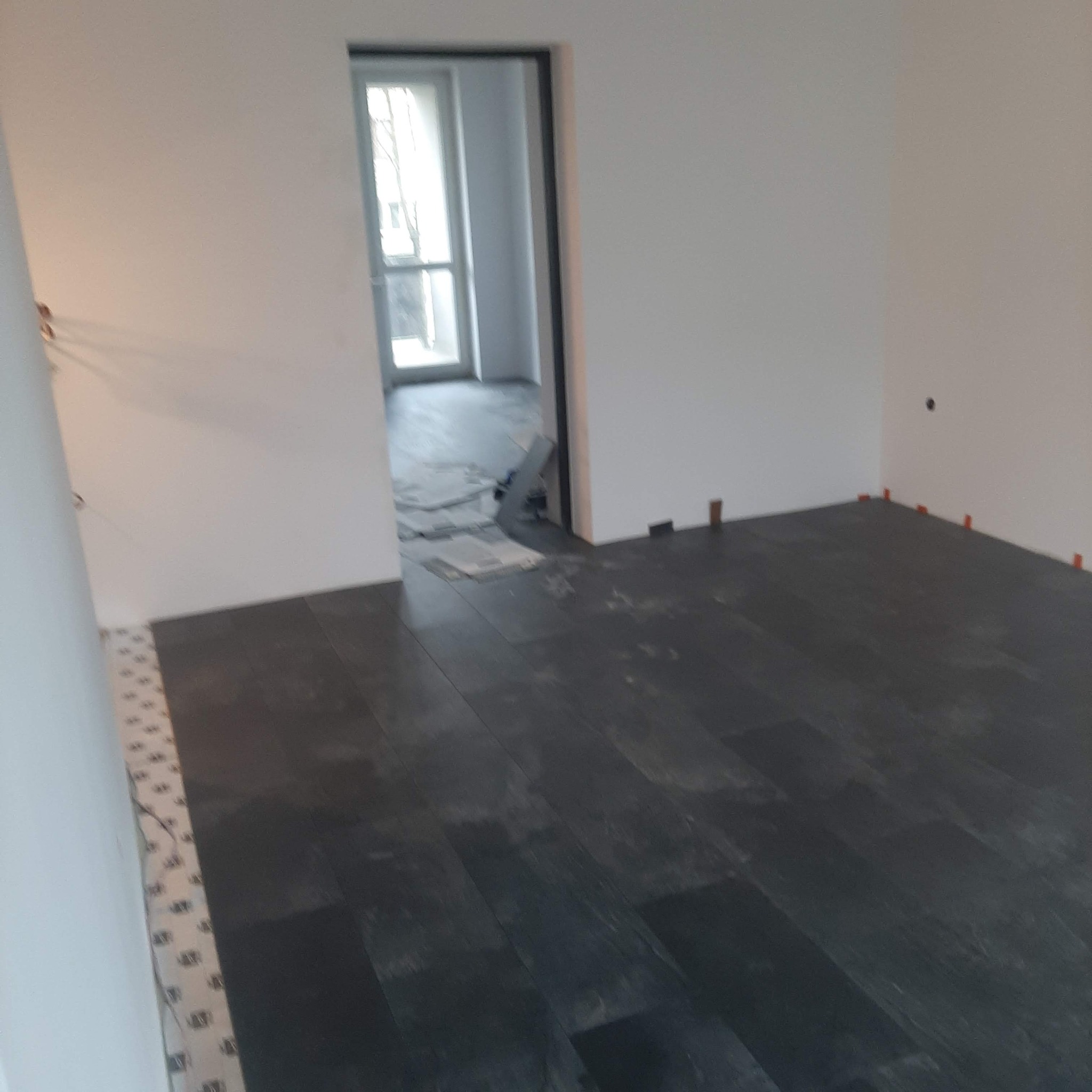 Układanie ciemnej podłogi z płytek imitujących drewno w trakcie remontu, widoczne krzyżyki dystansowe i fragment ułożonej mozaiki przy ścianie.