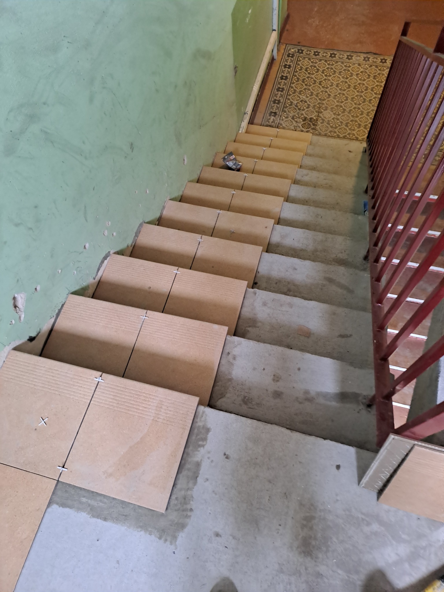 Montaż płytek na schodach w trakcie remontu, widoczne krzyżyki dystansowe, surowa ściana i czerwona barierka.