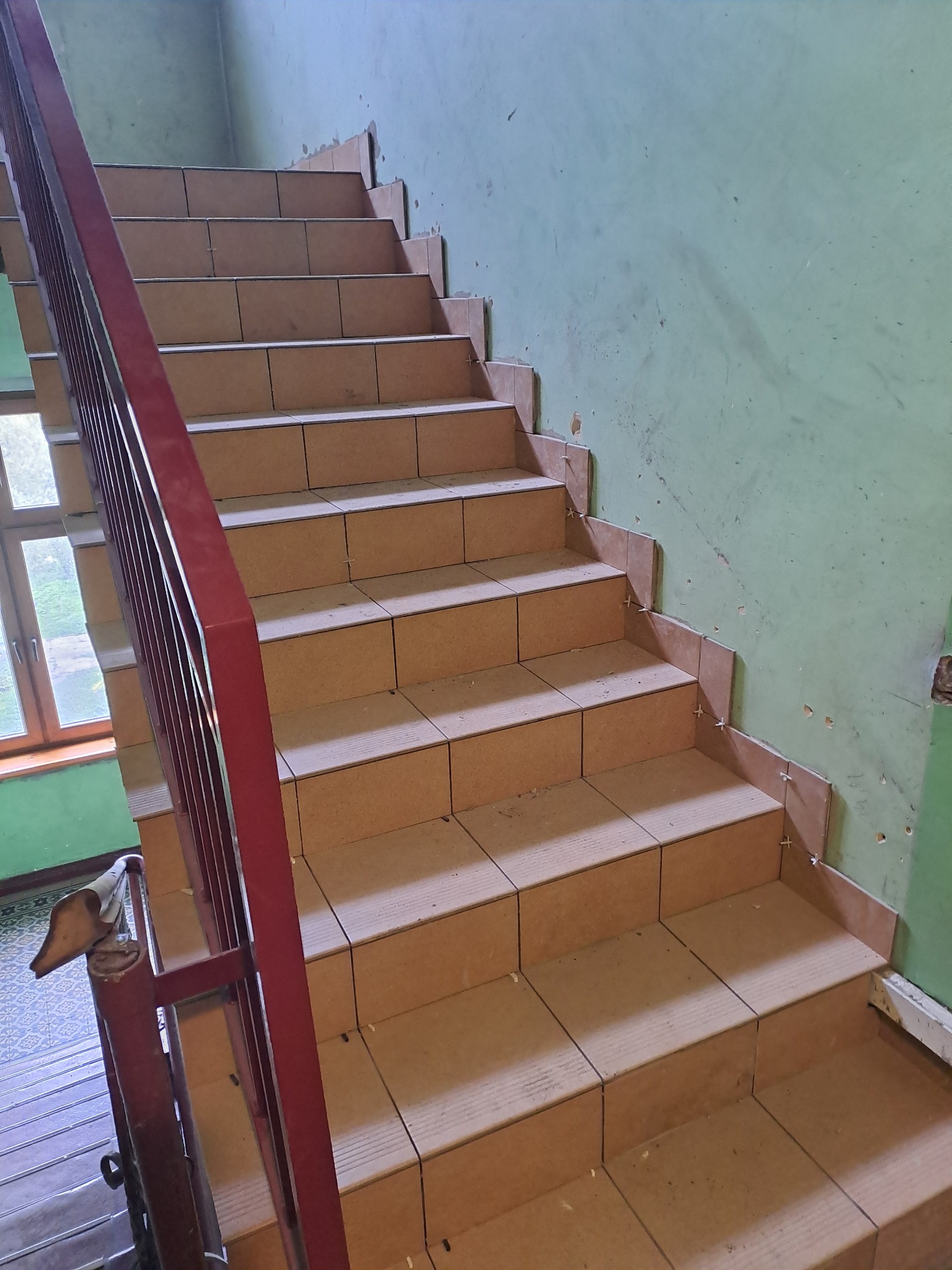 Klatka schodowa w trakcie układania płytek na stopniach, widoczne krzyżyki dystansowe, ściany w odcieniach zieleni z widocznymi śladami po pracach remontowych, czerwona metalowa balustrada.