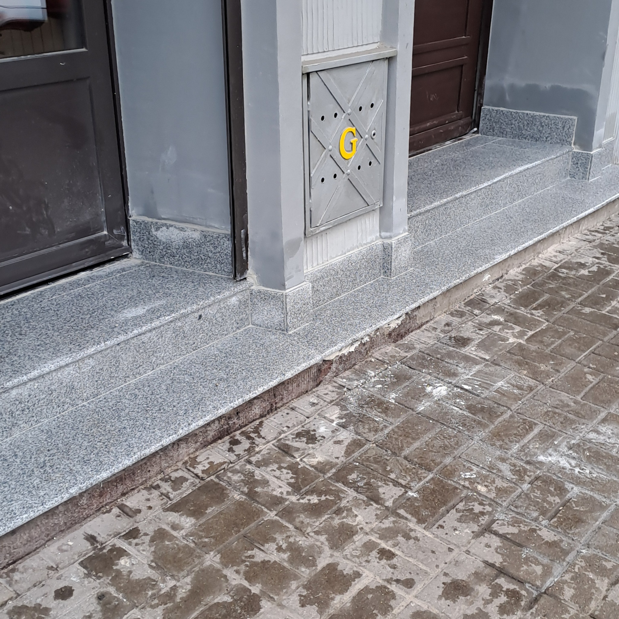 Wykończenie schodów wejściowych do budynku z szarego granitu, widoczna skrzynka na listy z żółtą literą G, mokra kostka brukowa przed wejściem.