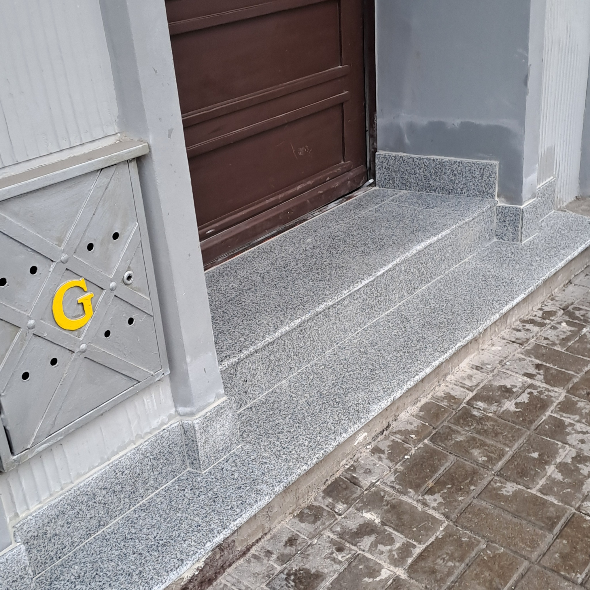 Wykończone granitowe schody wejściowe do budynku z brązowymi drzwiami, szara ściana z metalową skrzynką na listy z żółtą literą 'G'.