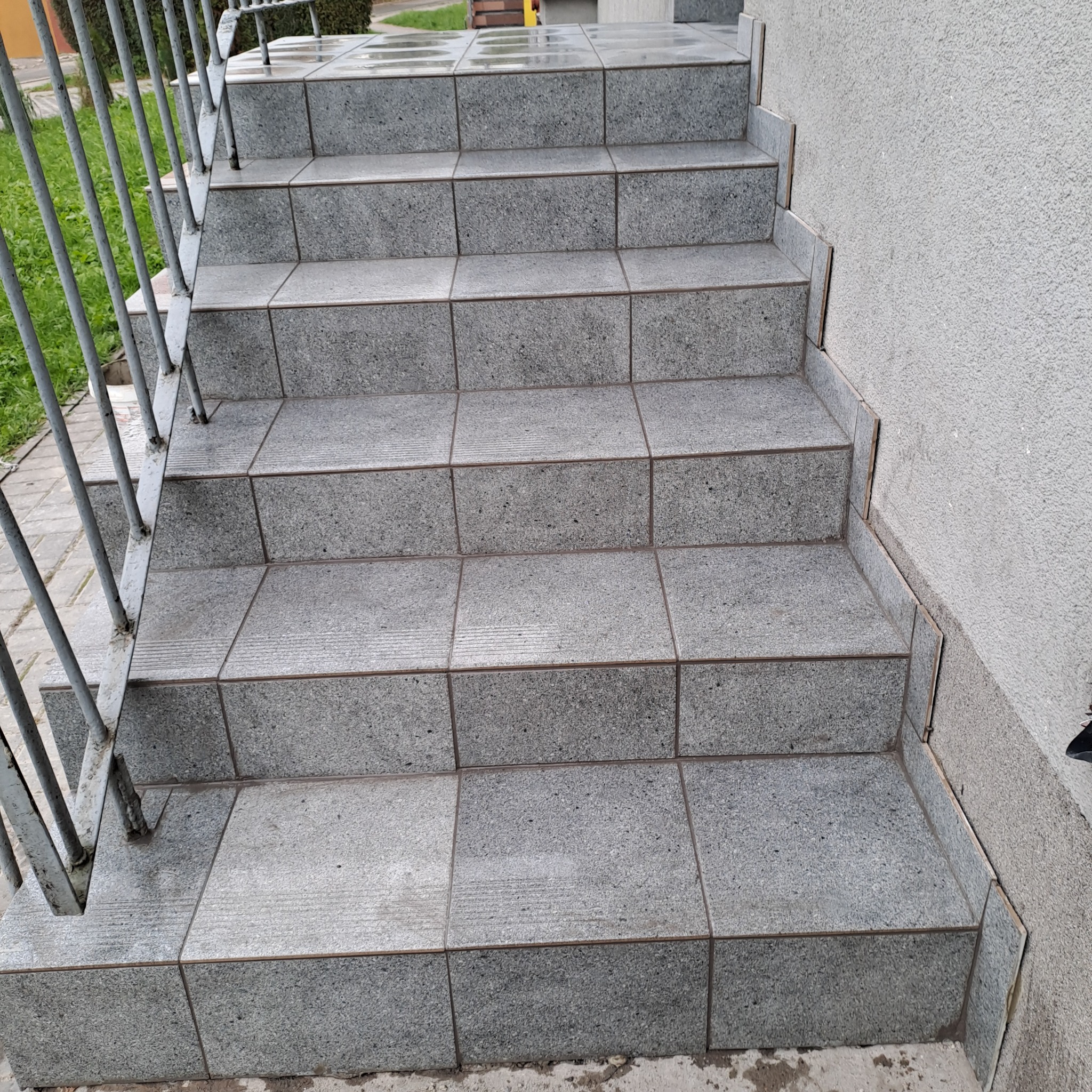 Zewnętrzne schody wejściowe wyłożone szarą, kwadratową płytką z delikatnym wzorem, zabezpieczone metalową, pomalowaną na szaro barierką; widoczne niewielkie zabrudzenia na krawędziach płytek.