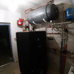 Czarny piec grzewczy z instalacją hydrauliczną, zbiornikiem buforowym i naczyniami wzbiorczymi w kotłowni w Kazimierzu Dolnym, widoczne rury miedziane i plastikowe.