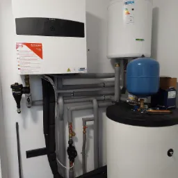 Instalacja grzewcza w kotłowni: biały zbiornik ciepłej wody, ekspansomat w kolorze niebieskim,  rury izolowane szarą pianką, czarny filtr TF1 Total Filter oraz pompa obiegowa Grundfos Alpha1 L...