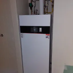 Biała pompa ciepła Viessmann z czarnym wyświetlaczem temperatury 10 stopni Celsjusza, zainstalowana w rogu łazienki z widocznymi rurami instalacji grzewczej.