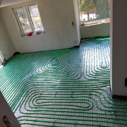 Zielone rury grzewcze ułożone spiralnie na folii izolacyjnej w jasnym, surowym pomieszczeniu z dwoma oknami; widoczne kable wystające ze ściany.