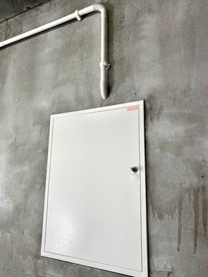 Biała skrzynka zamykana na kluczyk, zamontowana na szarej, surowej ścianie betonowej, z widocznymi rurami instalacyjnymi u góry.