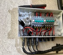 Otwarta skrzynka elektryczna z okablowaniem fotowoltaicznym, widoczne bezpieczniki, złącza MC4 i fragment narzędzia (szczypce) u góry.