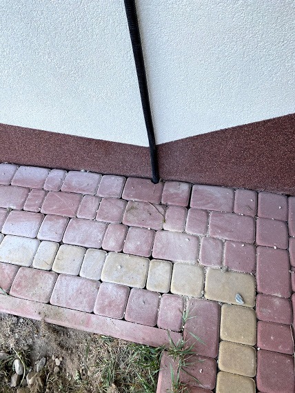 Czarna karbowana rura ochronna przymocowana do ściany budynku, biegnąca wzdłuż elewacji na tle kostki brukowej i fragmentów trawy.