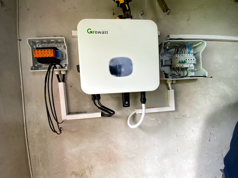 Biały falownik marki Growatt zamontowany na szarej ścianie, z widocznymi przewodami i puszkami elektrycznymi po bokach. Na górze wiertarka.