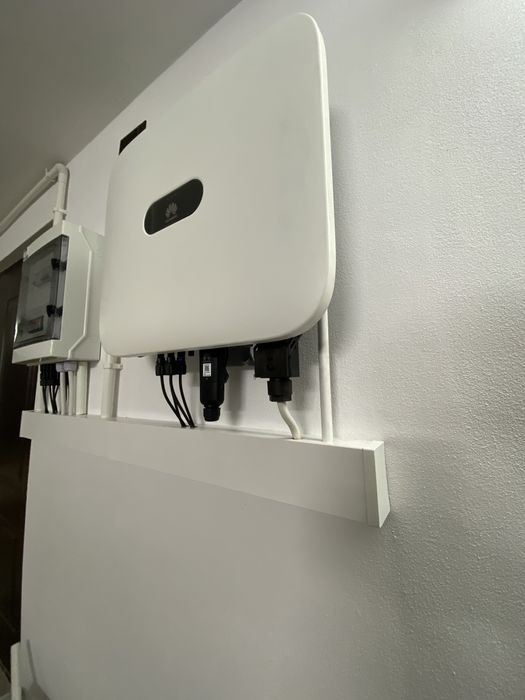 Biała skrzynka rozdzielcza i inwerter z logo na białej ścianie, widoczne kable i złącza, ujęcie z dołu.