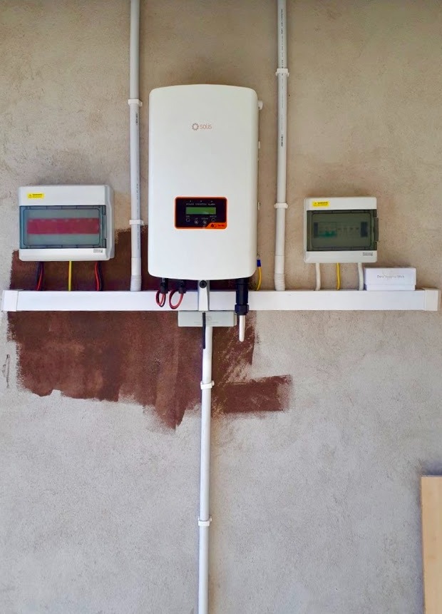 Biały falownik Solis zamontowany na szarej ścianie, z widocznymi przewodami i dwiema skrzynkami rozdzielczymi, podłączenie instalacji w trakcie prac wykończeniowych na ścianie.