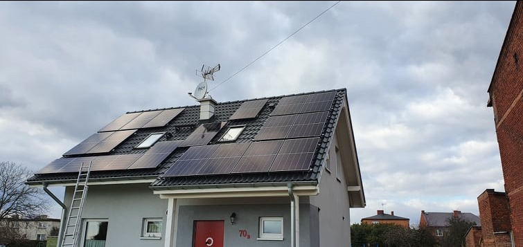 Dom jednorodzinny z panelami słonecznymi na dachu, drabina oparta o ścianę, pochmurne niebo w tle.