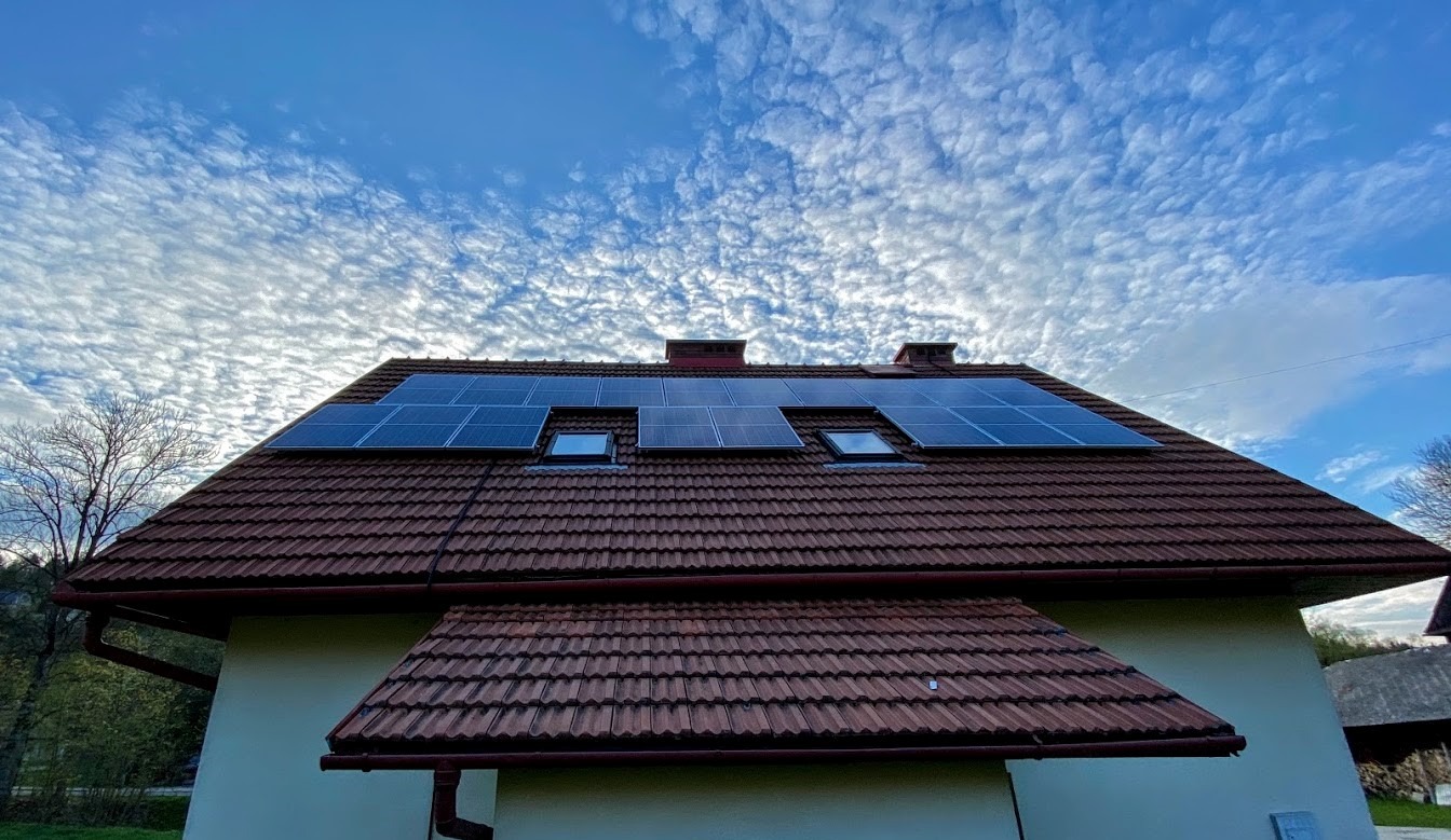 Panele słoneczne na dachu domu z dachówką w kolorze ceglastym, widok z dołu na tle błękitnego nieba z chmurami pierzastymi.
